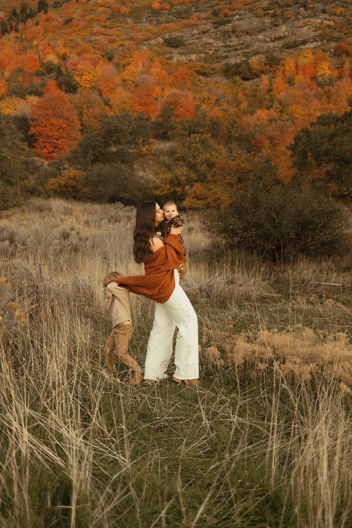 Mother holding her baby while her toddler pulls on her sweater in a beautiful Utah field with fall foliage.